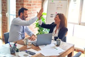 Woman and man, 40s, in business attire high five over laptop and desk covered in papers, with coffee cups in office with window, plant and exposed brick.