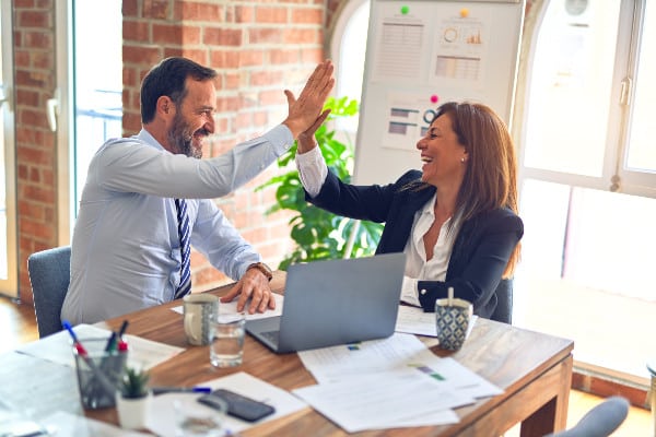 Woman and man, 40s, in business attire high five over laptop and desk covered in papers, with coffee cups in office with window, plant and exposed brick.