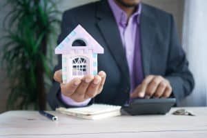 Diverse professional, man in dark suit with purple shirt holding toy house facade in outstretched hand, other hand on calculator with notebook and pen