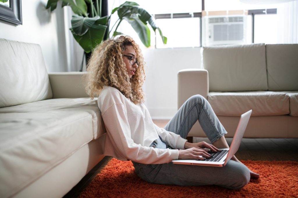 Woman in profile sitting on floor with computer on lap in living room with red carpet and white couches, entrepreneur