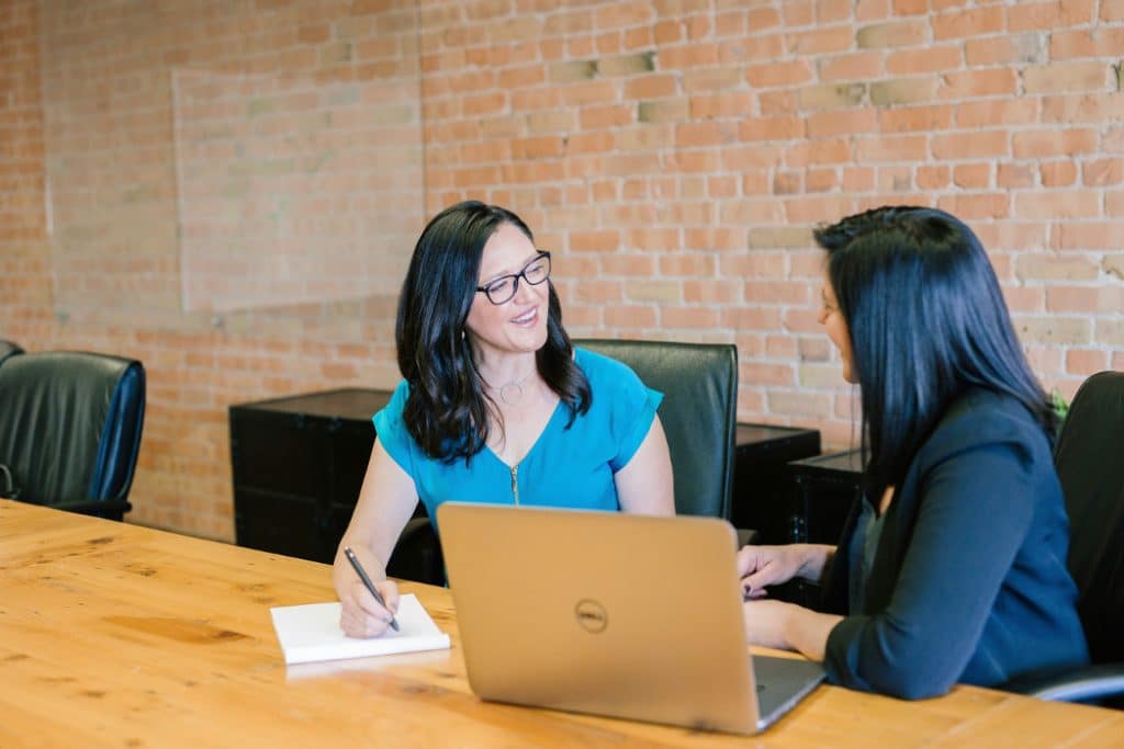 Dark haired women at boardroom table with brick wall background discussion with pen and paper and laptop, high net worth divorce mediation consultation
