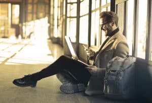 Man in business casual clothes sitting in airport with laptop and bags smiling by windows