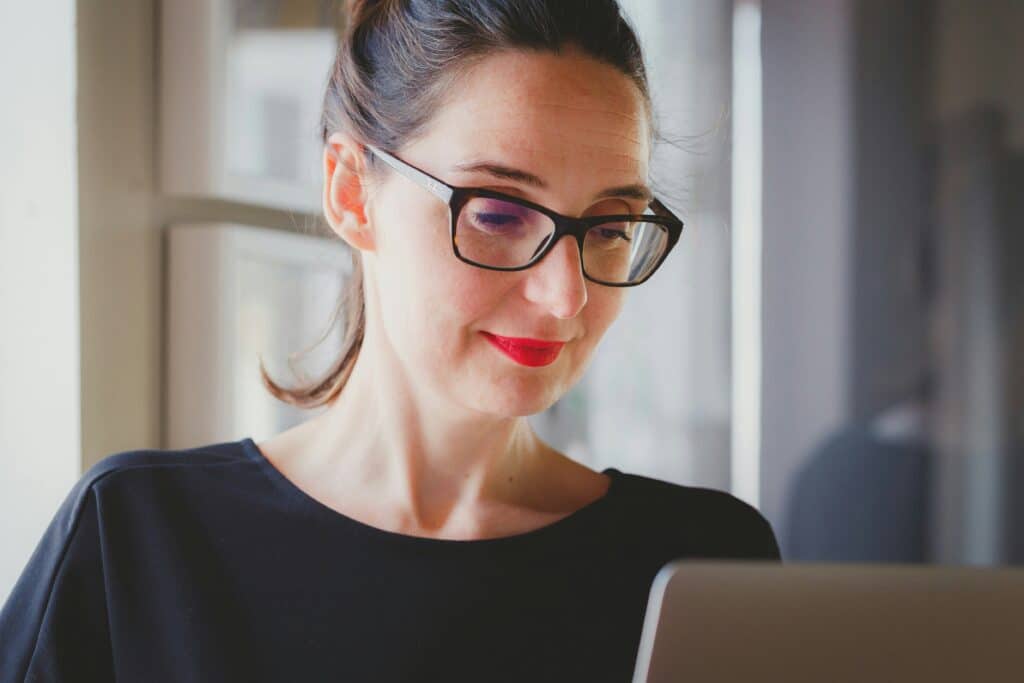 Woman in smart business attire and glasses smiles while looking at laptop in serene room