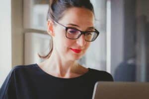 Woman in smart business attire and glasses smiles while looking at laptop in serene room